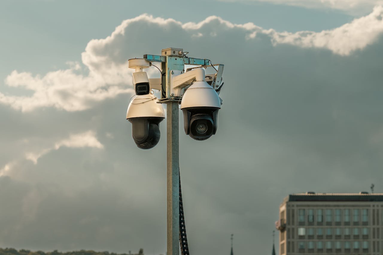 About Two modern surveillance cameras mounted on a pole in an urban setting under a cloudy sky.